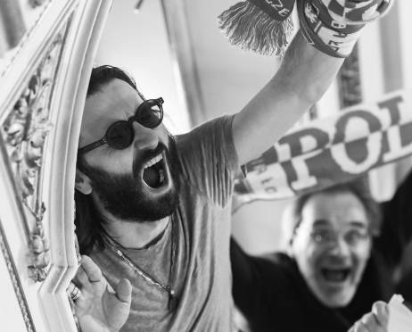 Czarno-białe zdjęcie. Mężczyzna wychyla się z przejścia, z uniesioną pięścią i otwartymi ustami, jakby skandował hasło. Ma na sobie ciemne okulary przeciwsłoneczne, a wokół nadgarstka owinięty szalik. W tle widoczna jest druga osoba, przejęta i zaangażowana.